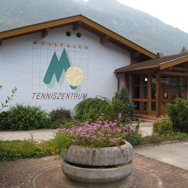 Ein Gebäude mit der Aufschrift "Mölltaler Tenniszentrum" und einem großen Wandgemälde zum Thema Tennis an der Außenwand; im Vordergrund Blumen und Grünpflanzen.