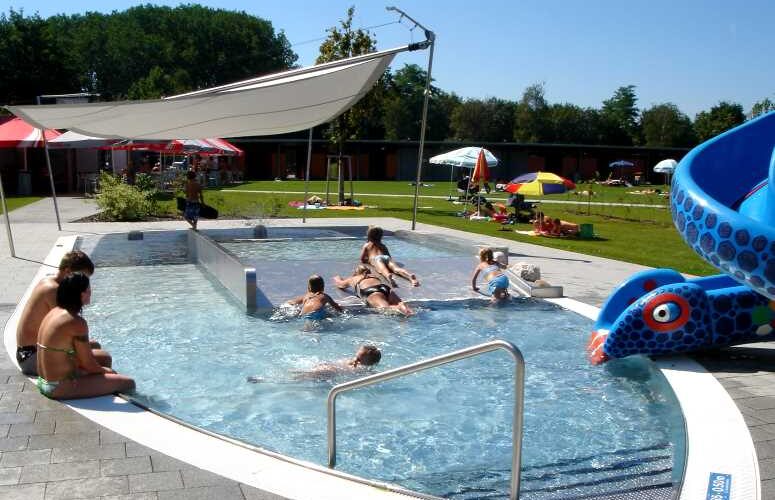 Kinder und Erwachsene schwimmen und spielen in der Nähe einer blauen Rutsche in einem Außenpool mit Sonnenschirmen und Rasenflächen.