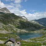 Eine Berglandschaft mit einem kleinen See, felsigen grünen Hügeln, ein paar Schneeflecken, einer Hütte im Vordergrund und einem teilweise bewölkten Himmel.