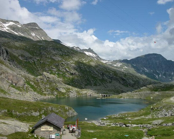 Eine Berglandschaft mit einem kleinen See, felsigen grünen Hügeln, ein paar Schneeflecken, einer Hütte im Vordergrund und einem teilweise bewölkten Himmel.