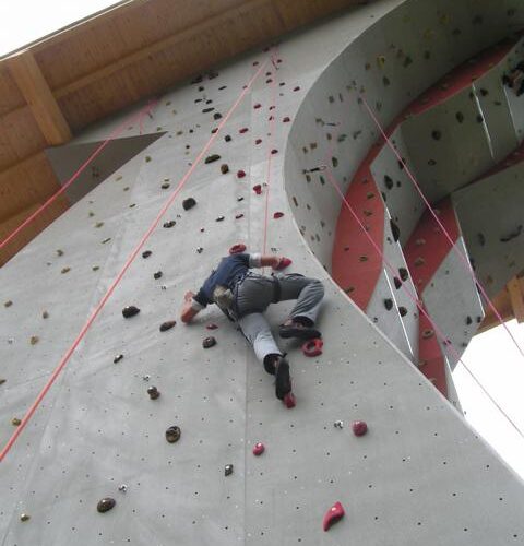 Eine Person klettert an einer künstlichen Felswand im Freien mit Hilfe von Griffen und Tritten, wobei ein Sicherungsseil über dem Kopf angebracht ist.