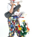 Ein Clown in bunten Patchworkhosen und einem hohen Hut spielt Flöte und hält einen Geigenkasten in der Hand. Er steht neben einer Topfpflanze mit künstlichen Blumen und einem Notenständer.