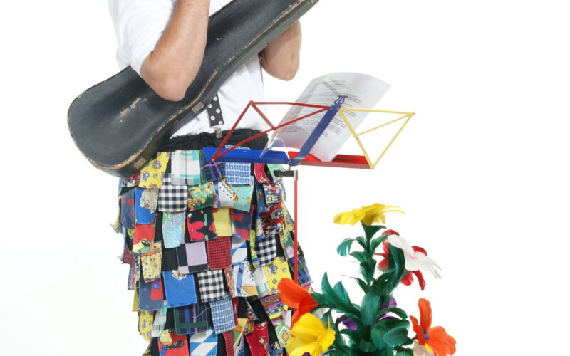 Ein Clown in bunten Patchworkhosen und einem hohen Hut spielt Flöte und hält einen Geigenkasten in der Hand. Er steht neben einer Topfpflanze mit künstlichen Blumen und einem Notenständer.