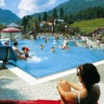 Menschen schwimmen und spielen in einem großen Freibad, umgeben von Bergen und Bäumen, während eine Frau am Rand sitzt und die Szene beobachtet.