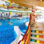 Eine Frau in einem roten Bikini steht auf einer bunt gefliesten Treppe mit Blick auf ein Hallenbad, in dem Kinder im Wasser spielen.