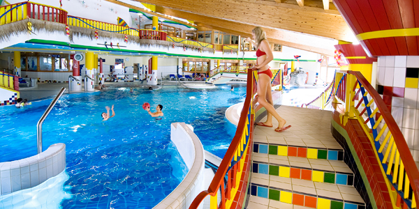 Eine Frau in einem roten Bikini steht auf einer bunt gefliesten Treppe mit Blick auf ein Hallenbad, in dem Kinder im Wasser spielen.