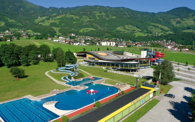 Ein großer Freibadkomplex mit Rutschen und einem modernen Gebäude, umgeben von grünen Feldern, Bäumen und einer Kulisse aus bewaldeten Hügeln und einer kleinen Stadt.