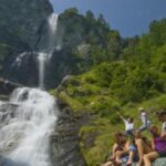 Eine Gruppe von Menschen sitzt auf grasbewachsenen Felsen in der Nähe des Fußes eines hohen Wasserfalls, umgeben von grünen Bäumen und steilen Felsen unter blauem Himmel.