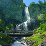 Eine Gruppe von Menschen steht auf einer Holzbrücke über einen Bach, mit einem hohen Wasserfall und felsigen Klippen umgeben von Grün im Hintergrund.