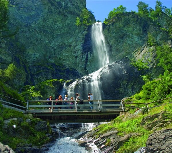 Eine Gruppe von Menschen steht auf einer Holzbrücke über einen Bach, mit einem hohen Wasserfall und felsigen Klippen umgeben von Grün im Hintergrund.