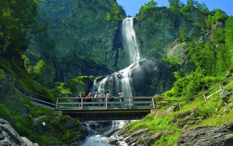 Eine Gruppe von Menschen steht auf einer Holzbrücke über einen Bach, mit einem hohen Wasserfall und felsigen Klippen umgeben von Grün im Hintergrund.
