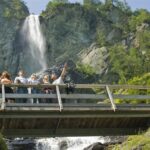 Eine Gruppe von Menschen steht auf einer Holzbrücke und macht ein Selfie, mit einem Wasserfall und grünen Bäumen im Hintergrund.