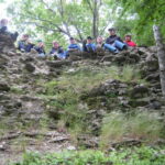 Eine Gruppe von Menschen sitzt am Rande einer felsigen Klippe und blickt nach unten, umgeben von Bäumen und Grünzeug.