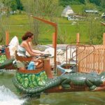 Zwei Personen fahren auf einer kleinen Wasserattraktion in einem Vergnügungspark mit Booten in Tierform, die wie Krokodile geformt sind, mit einer Giraffenstatue und einem eingezäunten Bereich im Hintergrund.