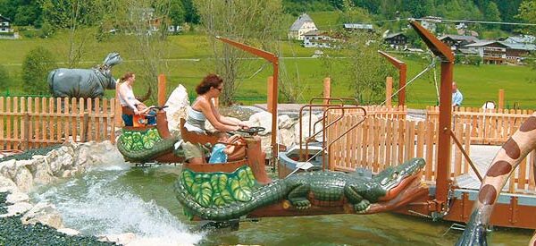 Zwei Personen fahren auf einer kleinen Wasserattraktion in einem Vergnügungspark mit Booten in Tierform, die wie Krokodile geformt sind, mit einer Giraffenstatue und einem eingezäunten Bereich im Hintergrund.