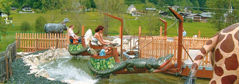Zwei Personen fahren auf einer kleinen Wasserattraktion in einem Vergnügungspark mit Booten in Tierform, die wie Krokodile geformt sind, mit einer Giraffenstatue und einem eingezäunten Bereich im Hintergrund.