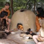 Vier Personen in historischer Kleidung backen Brot an einem Lehmofen im Freien, umgeben von Bäumen und belaubten Ästen.