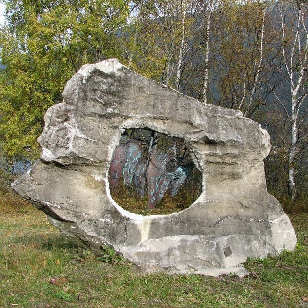 Ein großer, verwitterter Felsen mit einem natürlichen, kreisrunden Loch steht auf einem grasbewachsenen Boden, mit Bäumen und Hügeln im Hintergrund.