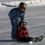 Zwei Kinder, die Helme und Winterkleidung tragen, laufen Schlittschuh; das eine steht und hilft dem anderen, das auf dem Eis sitzt.