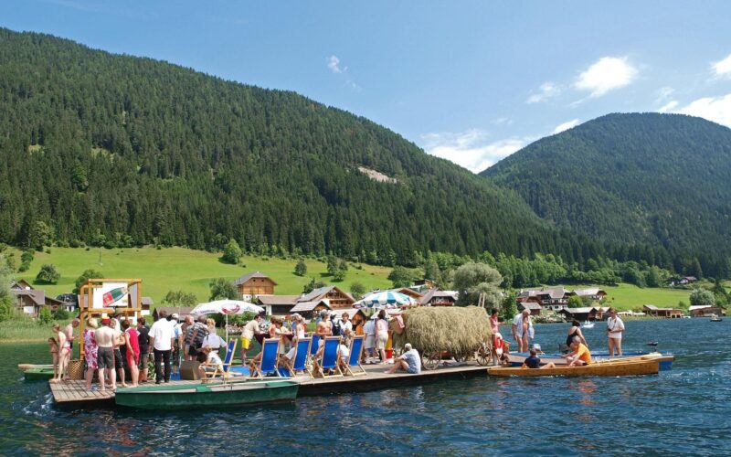 Eine Gruppe von Menschen steht und sitzt auf einem großen Holzfloß mit Liegestühlen und Sonnenschirmen auf einem See, umgeben von grünen Hügeln und Häusern.