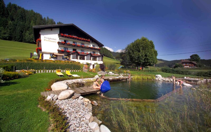 Ein Hotel mit blumengeschmückten Balkonen steht an einem natürlichen Schwimmteich, in dem sich die Menschen entspannen; grüne Wiesen und Hügel umgeben das Gebiet unter einem klaren blauen Himmel.