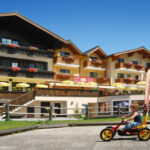 Ein Kind fährt mit einem Tret-Gokart auf einem gepflasterten Weg vor einem mehrstöckigen Hotel im alpinen Stil mit Balkonen und Blumenkästen.