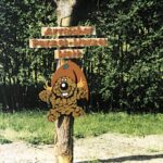 Ein hölzernes Schild mit deutschem Text und einer Cartoon-Biberfigur mit rotem Hut ist an einem Baum befestigt; im Hintergrund ist ein Wald zu sehen.