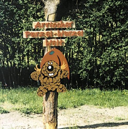 Ein hölzernes Schild mit deutschem Text und einer Cartoon-Biberfigur mit rotem Hut ist an einem Baum befestigt; im Hintergrund ist ein Wald zu sehen.