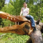 Eine junge Frau sitzt auf einem großen umgestürzten Baum, der wie ein Krokodilkopf mit geschnitzten Zähnen aussieht, in einem Waldgebiet.