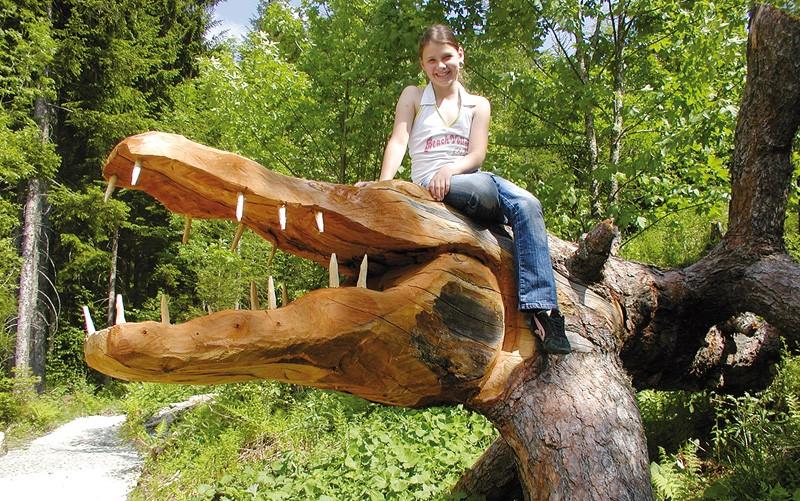 Eine junge Frau sitzt auf einem großen umgestürzten Baum, der wie ein Krokodilkopf mit geschnitzten Zähnen aussieht, in einem Waldgebiet.