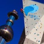 Eine Person klettert an einer künstlichen Kletterwand im Freien neben einem hohen, blau-silbernen Aussichtsturm vor einem klaren blauen Himmel.