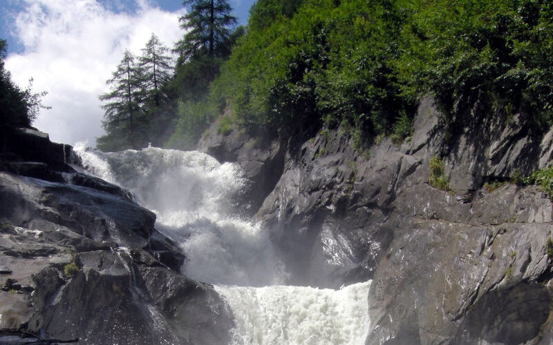 Ein mächtiger Wasserfall fließt über dunkle Felsklippen, umgeben von dichten grünen Bäumen und Vegetation unter einem teilweise bewölkten Himmel.