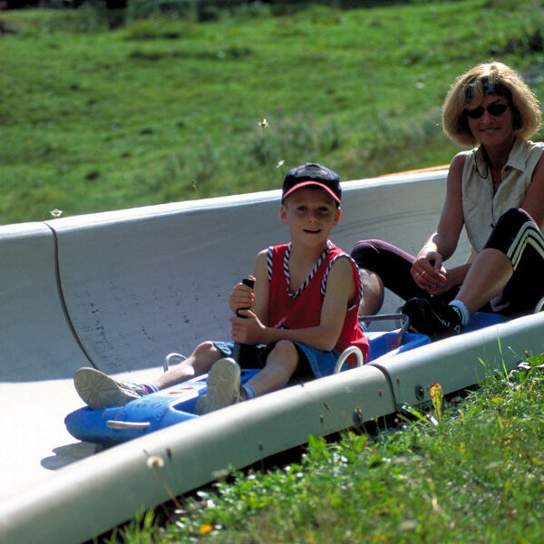 Eine Frau und ein Kind fahren gemeinsam in einem Zweipersonenschlitten eine Betonrodelbahn auf einem grasbewachsenen Hügel hinunter.