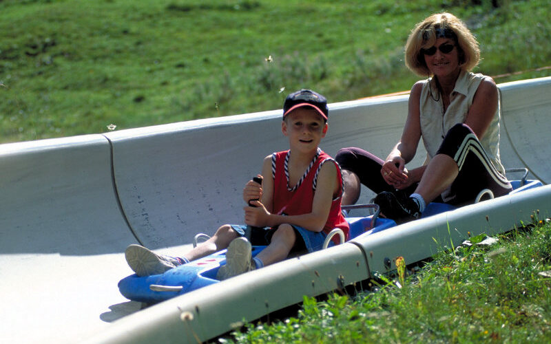 Eine Frau und ein Kind fahren gemeinsam in einem Zweipersonenschlitten eine Betonrodelbahn auf einem grasbewachsenen Hügel hinunter.