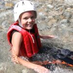 Ein Kind, das eine rote Schwimmweste und einen weißen Helm trägt, sitzt im flachen, klaren Wasser und lächelt in die Kamera.