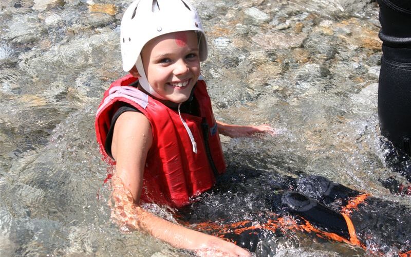 Ein Kind, das eine rote Schwimmweste und einen weißen Helm trägt, sitzt im flachen, klaren Wasser und lächelt in die Kamera.