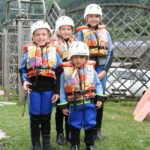 Vier Kinder, die Neoprenanzüge, bunte Schwimmwesten und Helme tragen, stehen im Freien zusammen, während rechts die Paddel zu sehen sind.