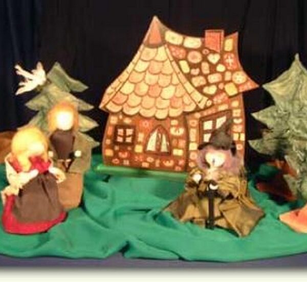Drei Puppen und zwei Bäume sind vor einer Lebkuchenhaus-Kulisse auf einem grünen Tuch angeordnet, die eine Szene aus Hänsel und Gretel" darstellt.