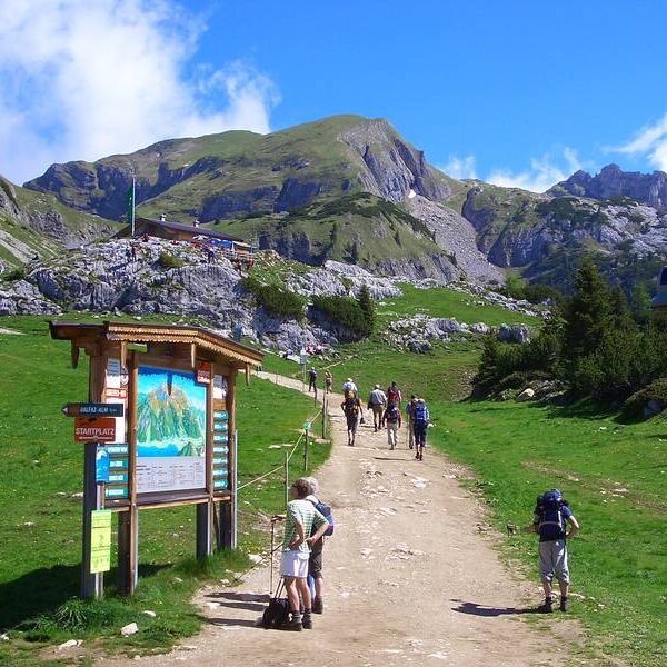 Wanderer gehen einen breiten unbefestigten Weg in einer bergigen Landschaft hinauf, mit einem großen Schild im Vordergrund und grünen Hügeln und felsigen Gipfeln im Hintergrund.