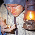 Eine Person mit Helm und warmer Kleidung untersucht im Licht einer Laterne einen Goldnugget, wobei Gesicht und Hände mit Staub bedeckt sind.