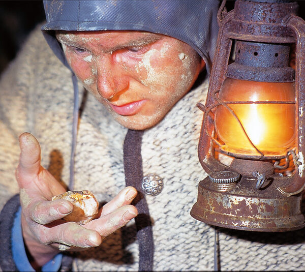 Eine Person mit Helm und warmer Kleidung untersucht im Licht einer Laterne einen Goldnugget, wobei Gesicht und Hände mit Staub bedeckt sind.