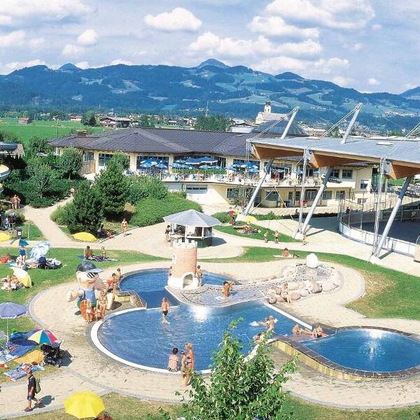 Außenpoolkomplex mit Menschen, die sich sonnen und schwimmen, umgeben von Sonnenschirmen, Gras und Gebäuden, im Hintergrund sind Berge zu sehen.