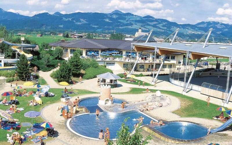 Außenpoolkomplex mit Menschen, die sich sonnen und schwimmen, umgeben von Sonnenschirmen, Gras und Gebäuden, im Hintergrund sind Berge zu sehen.