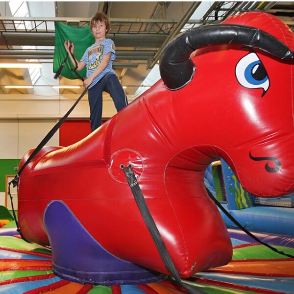 Ein Kind reitet auf einem roten, aufblasbaren mechanischen Stier in einem bunt geschmückten Innenspielbereich.