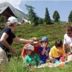 Eine Gruppe von Erwachsenen und Kindern macht ein Picknick auf einem grasbewachsenen Hügel mit Bäumen, Bergen und einer Hütte im Hintergrund.