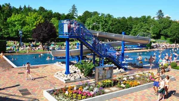 Öffentliches Freibad mit blauer Wasserrutsche, umgeben von Bäumen und Menschen, die an einem sonnigen Tag schwimmen, sich sonnen und spazieren gehen.