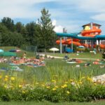 Menschen entspannen sich auf einer Wiese mit Handtüchern und Sonnenschirmen in der Nähe eines Wasserparks mit orangefarbenen und blauen Wasserrutschen, umgeben von Bäumen und Blumen.