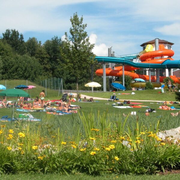 Menschen entspannen sich auf einer Wiese mit Handtüchern und Sonnenschirmen in der Nähe eines Wasserparks mit orangefarbenen und blauen Wasserrutschen, umgeben von Bäumen und Blumen.