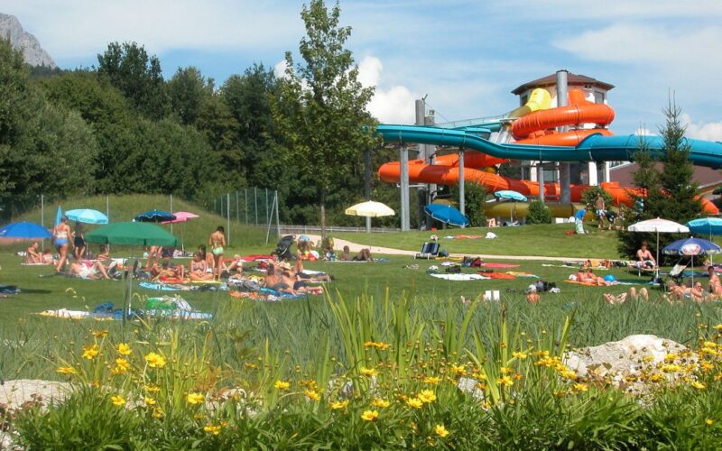 Menschen entspannen sich auf einer Wiese mit Handtüchern und Sonnenschirmen in der Nähe eines Wasserparks mit orangefarbenen und blauen Wasserrutschen, umgeben von Bäumen und Blumen.