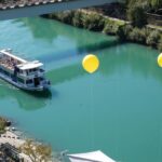 Ein Passagierboot fährt auf einem türkisfarbenen Fluss unter einer Brücke hindurch, in dessen Nähe mehrere gelbe Luftballons schwimmen und Bäume das Ufer säumen.